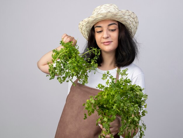 Jovem morena satisfeita com um jardineiro de uniforme, usando chapéu de jardinagem, segurando e olhando para o coentro isolado na parede branca