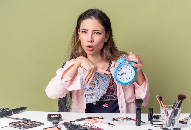 Jovem morena sem noção sentada à mesa com ferramentas de maquiagem segurando dinheiro e apontando para o despertador