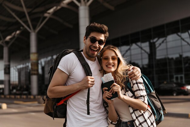 Foto grátis jovem morena sorri amplamente e abraça sua namorada atraente casal de viajantes segura mochilas e passaportes no fundo do aeroporto