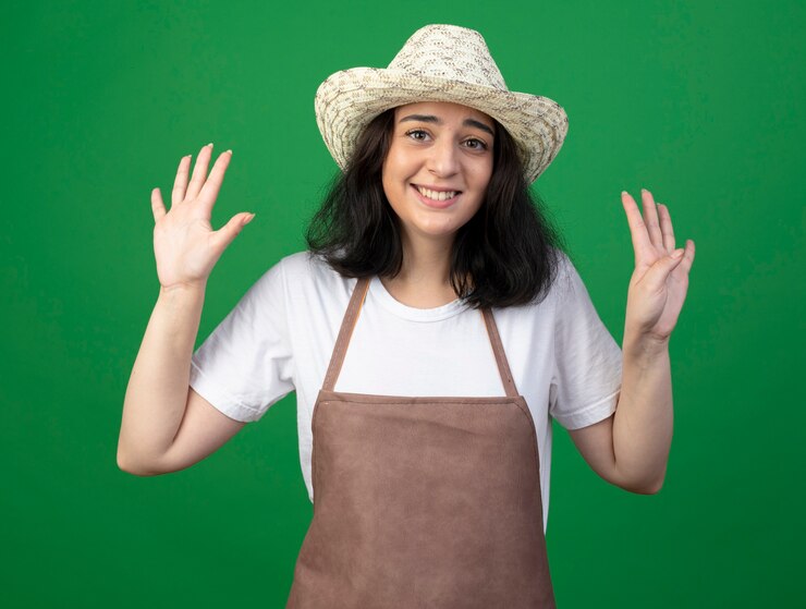 Foto grátis jovem morena sorridente, jardineira feminina, usando óculos óticos e uniforme, usando um chapéu de jardinagem, gestos nove com dedos isolados na parede verde