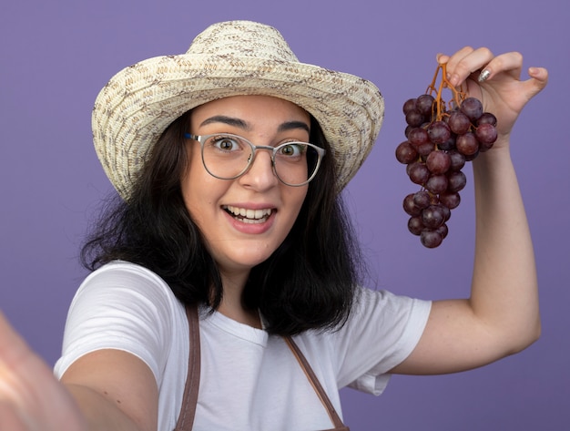 Jovem morena surpresa com óculos e uniforme, usando chapéu de jardinagem segurando uvas olhando para frente isoladas na parede roxa