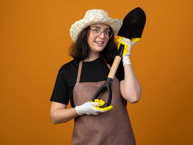 Jovem morena surpresa com óculos e uniforme, usando chapéu e luvas de jardinagem, segura a pá isolada na parede laranja