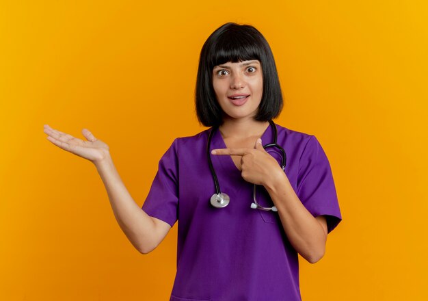 Jovem morena surpresa médica de uniforme com pontos de estetoscópio na mão vazia isolada em um fundo laranja com espaço de cópia