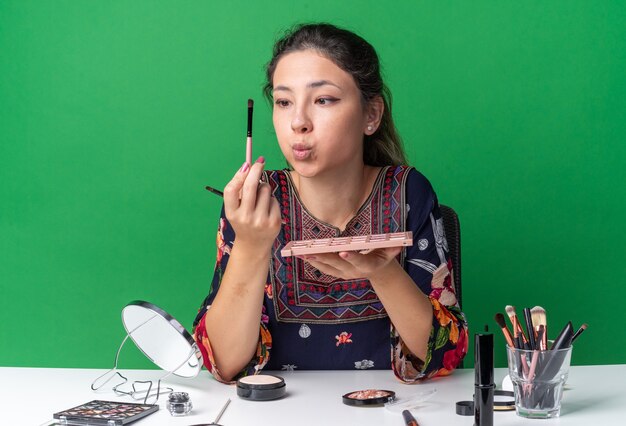 Jovem morena surpresa sentada à mesa com ferramentas de maquiagem segurando uma paleta de sombras e olhando para o pincel de maquiagem