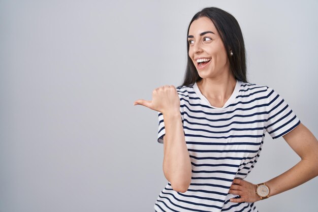 Jovem morena vestindo camiseta listrada sorrindo com cara de feliz olhando e apontando para o lado com o polegar para cima.