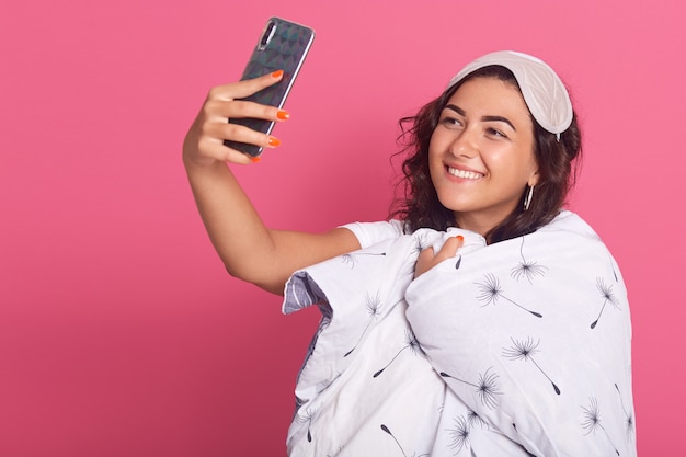 Foto grátis jovem morena vestindo máscara de dormir e embrulhado cobertor branco, tomando selfie durante a hora de dormir, garota se divertindo na manhã