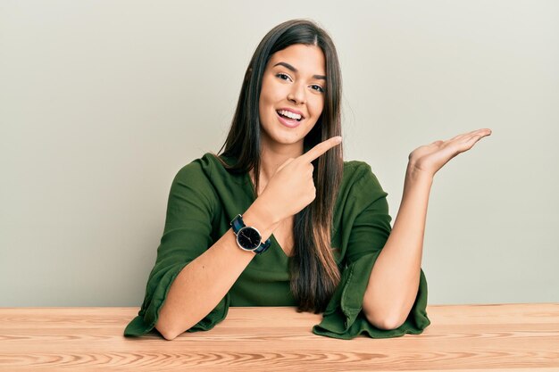 Jovem morena vestindo roupas casuais, sentada na mesa, maravilhada e sorrindo para a câmera enquanto apresenta a mão e aponta com o dedo.