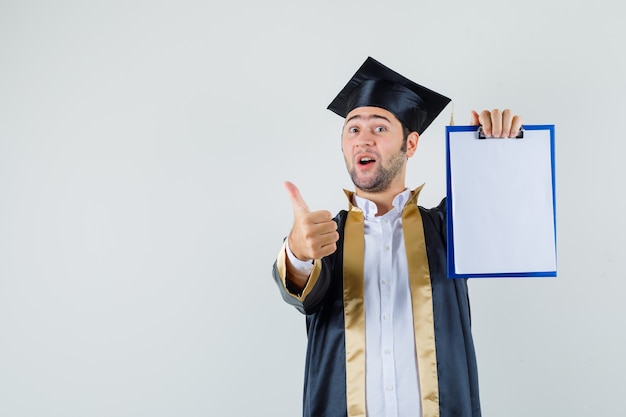 Jovem mostrando a prancheta com o polegar para cima em uniforme de pós-graduação e parecendo feliz. vista frontal.