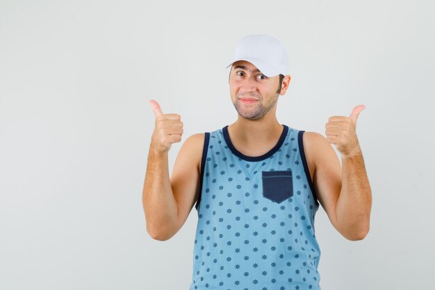 Jovem mostrando dois polegares para cima em camiseta azul, boné e olhando alegre, vista frontal.