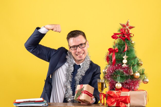 jovem mostrando músculo sentado à mesa perto da árvore de natal e presentes em amarelo