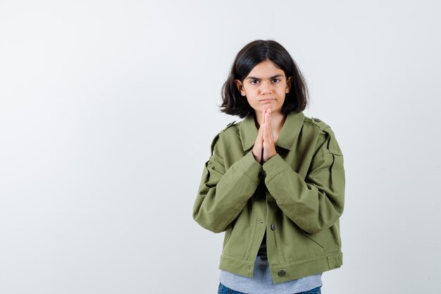 Jovem, mostrando o gesto namastê de suéter cinza, jaqueta cáqui, calça jeans e olhando séria, vista frontal.