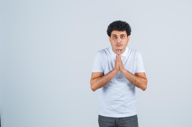 Jovem mostrando o gesto namastê em jeans e camiseta branca e olhando sério, vista frontal.