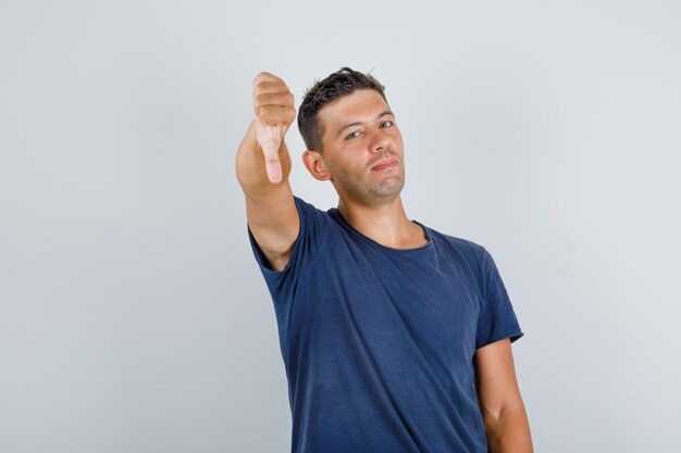 Jovem mostrando o polegar para baixo em uma camiseta azul escura e parecendo insatisfeito, vista frontal.