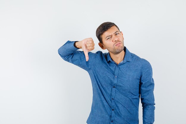 Jovem mostrando o polegar para baixo na camisa azul e parecendo insatisfeito. vista frontal.