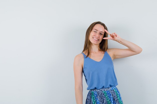 Jovem, mostrando o sinal de V na camiseta, saia e parecendo feliz, vista frontal.