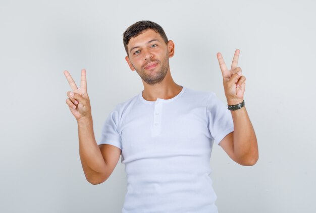 Jovem mostrando os dedos fazendo o sinal da vitória com uma camiseta branca e parecendo feliz, vista frontal