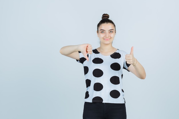 Jovem, mostrando os polegares para cima e para baixo em t-shirt, jeans e parecendo feliz. vista frontal.