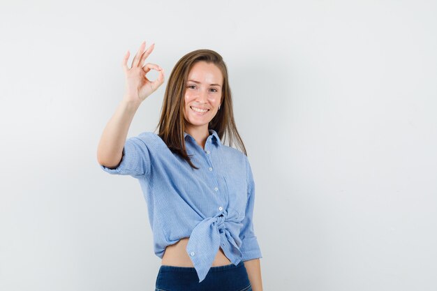 Jovem mostrando sinal de ok na camisa azul, calça e parecendo feliz.