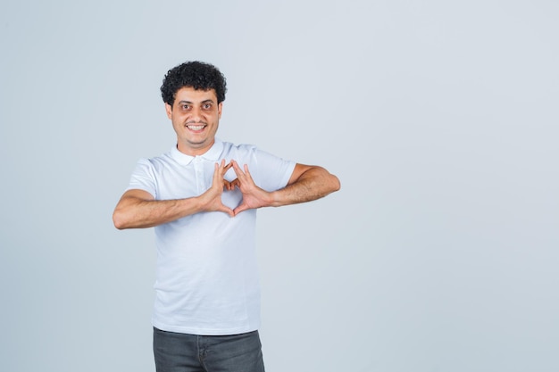 Jovem mostrando um gesto de coração em t-shirt branca, calças e olhando alegre, vista frontal.