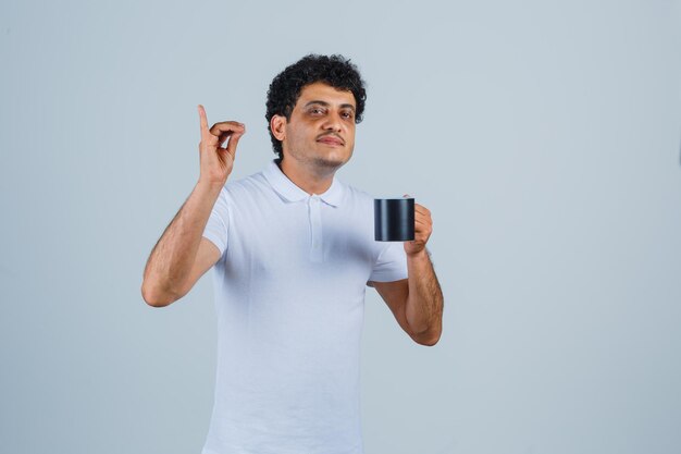 Jovem mostrando um gesto delicioso e segurando uma xícara de chá em jeans e camiseta branca e olhando feliz, vista frontal.