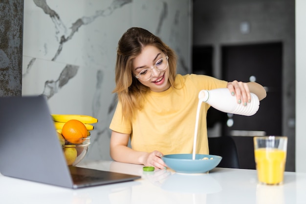 Jovem muito alegre tomando café da manhã enquanto está sentado à mesa da cozinha, trabalhando no laptop