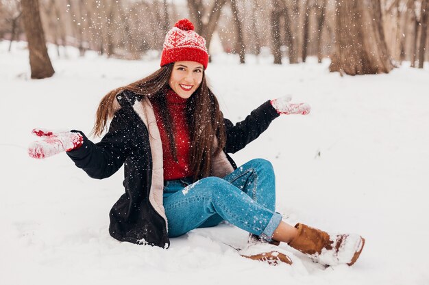 Jovem muito cândida, sorridente, feliz, com luvas vermelhas e chapéu de malha, vestindo um casaco preto, andando brincando no parque na neve, roupas quentes, se divertindo