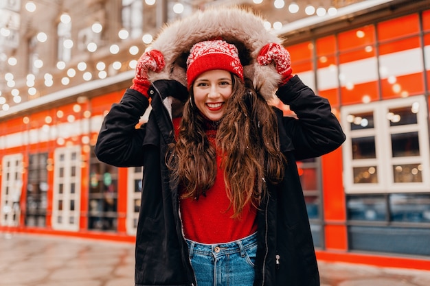 Jovem, muito sorridente, animada, mulher feliz com luvas vermelhas e chapéu de malha, vestindo um casaco de inverno, andando na rua da cidade, roupas quentes