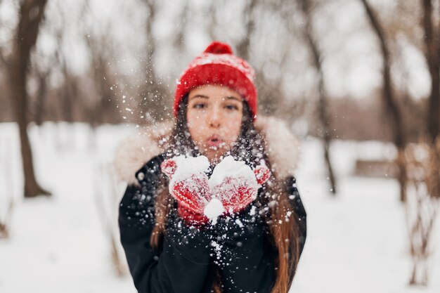 Jovem, muito sorridente, feliz, com luvas vermelhas e chapéu de malha, vestindo um casaco de inverno, caminhando no parque, soprando neve