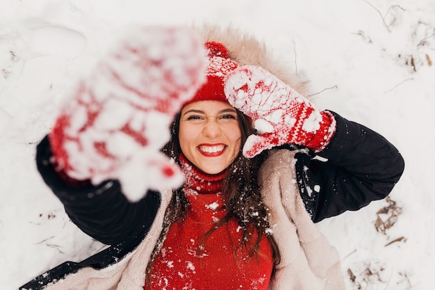 Jovem, muito sorridente, feliz, com luvas vermelhas e chapéu de malha, vestindo um casaco de inverno, deitado no parque na neve, roupas quentes, vista de cima