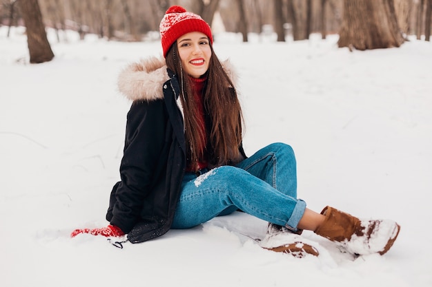 Jovem, muito sorridente, feliz, com luvas vermelhas e chapéu de malha, vestindo um casaco de inverno, sentado na neve no parque, roupas quentes