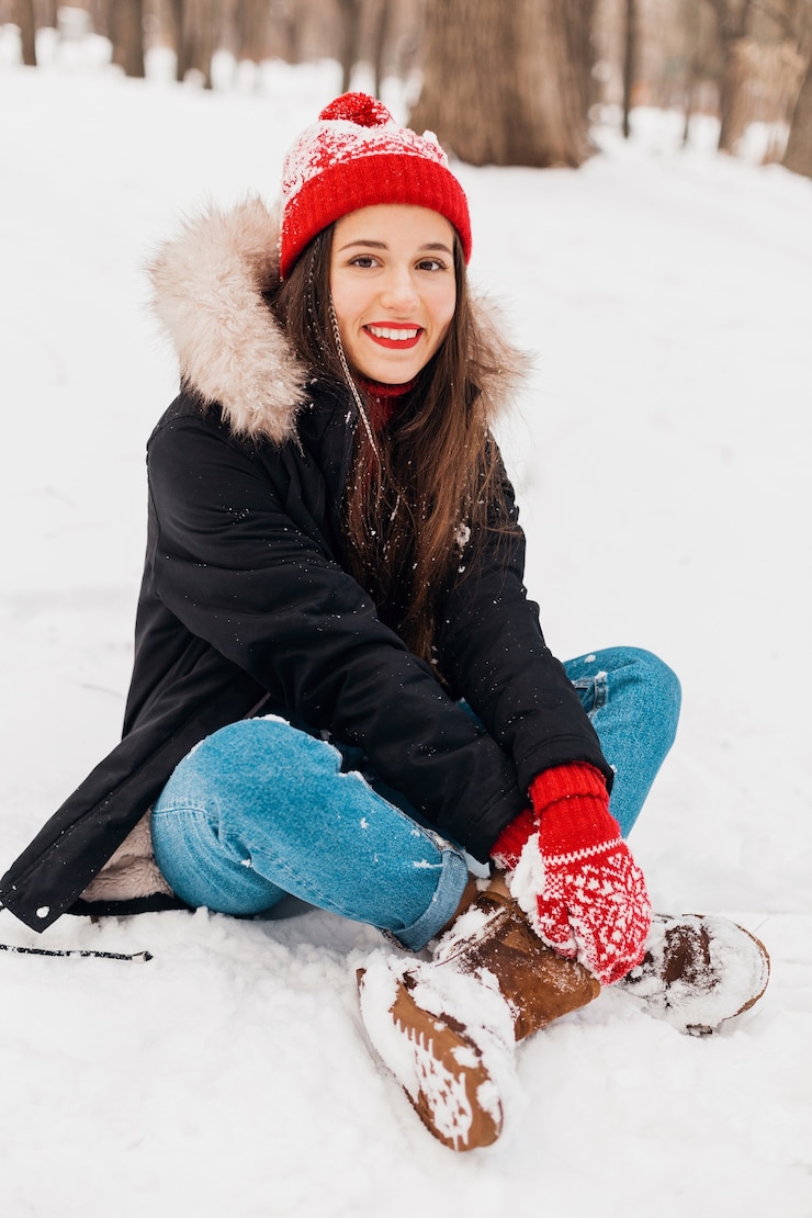 Foto grátis jovem, muito sorridente, feliz, com luvas vermelhas e chapéu de malha, vestindo um casaco de inverno, sentado na neve no parque, roupas quentes