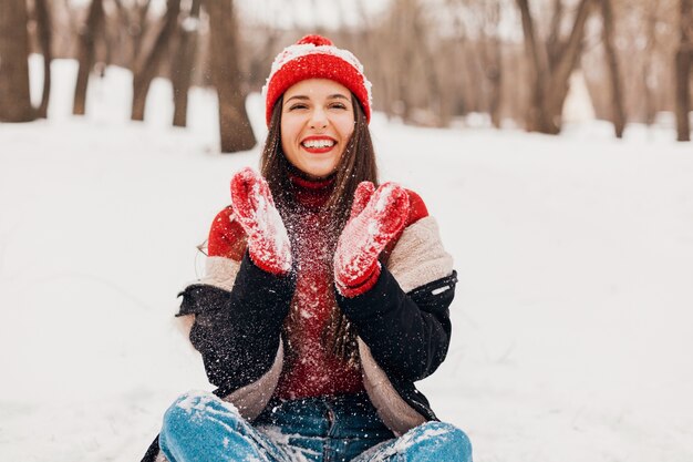 Jovem, muito sorridente, feliz, com luvas vermelhas e chapéu de malha, vestindo um casaco de inverno, sentado na neve no parque, roupas quentes