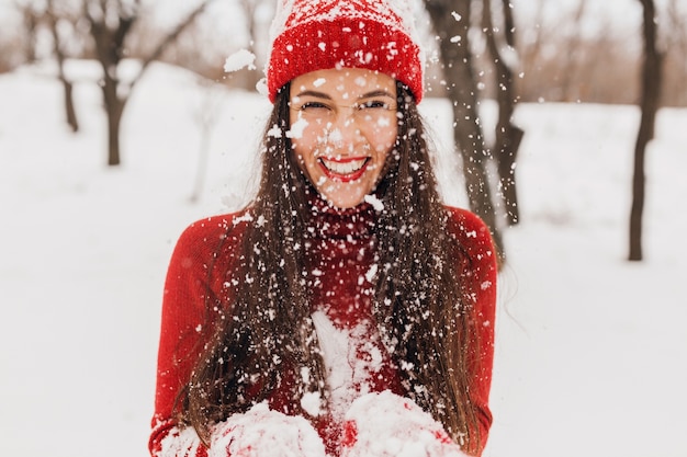 Jovem, muito sorridente, feliz, com luvas vermelhas e chapéu, suéter de malha, caminhando no parque na neve, roupas quentes, se divertindo