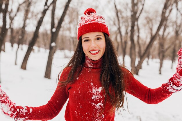 Jovem, muito sorridente, feliz, com luvas vermelhas e um chapéu, um suéter de malha andando no parque na neve, roupas quentes, se divertindo, balançando cabelos longos