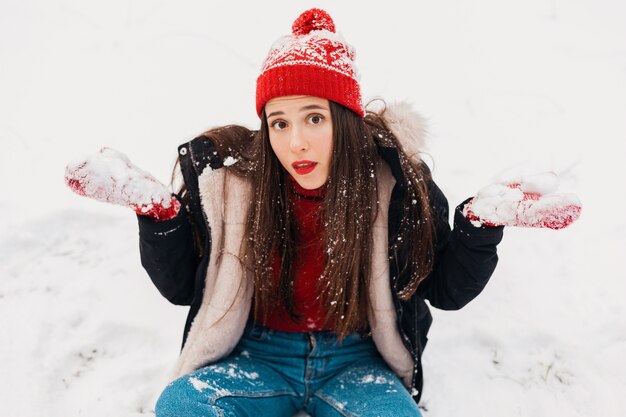 Jovem, muito sorridente, feliz, usando luvas vermelhas e chapéu de malha, vestindo um casaco de inverno, caminhando no parque, brincando com a neve em roupas quentes