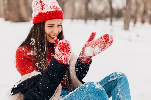 Jovem, muito sorridente, feliz, usando luvas vermelhas e chapéu de malha, vestindo um casaco de inverno, caminhando no parque, brincando com a neve em roupas quentes
