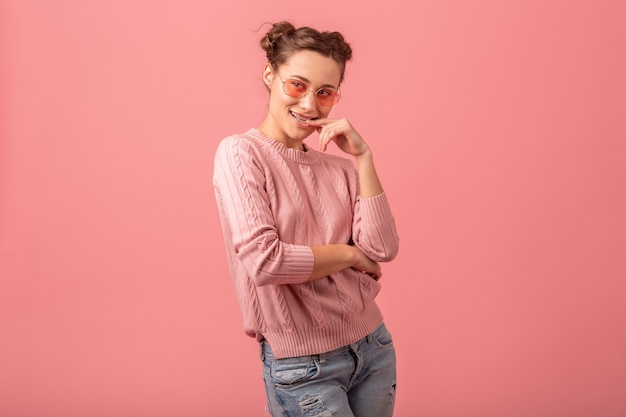 Jovem, muito sorridente, glamourosa, de suéter rosa e óculos de sol, isolados no fundo rosa do estúdio