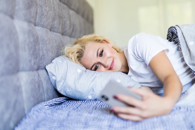 Jovem mulher acordando em sua cama e tomando seu smartphone, ela está verificando mensagens e cochilando alarme