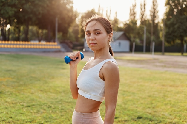 Jovem mulher adulta com aparência agradável, vestindo blusa esporte branca malhando bíceps e tríceps, usando halteres azuis para treinamento ao ar livre no estádio.