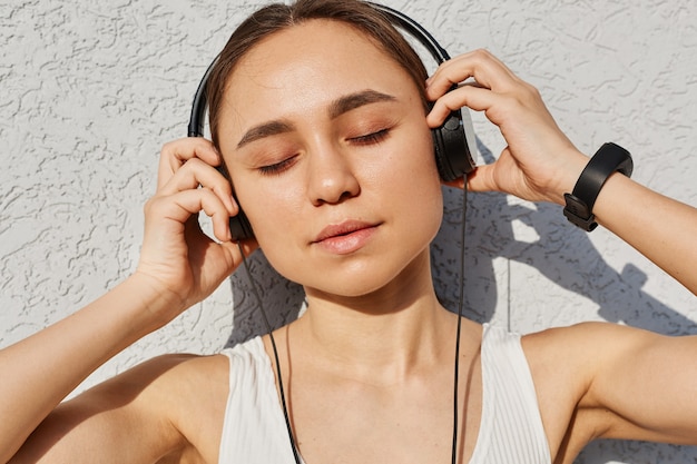 Jovem mulher adulta com cabelo escuro, blusa branca, mantendo os olhos fechados, tocando os fones de ouvido com as palmas das mãos, curtindo música depois do treino, estilo de vida saudável.