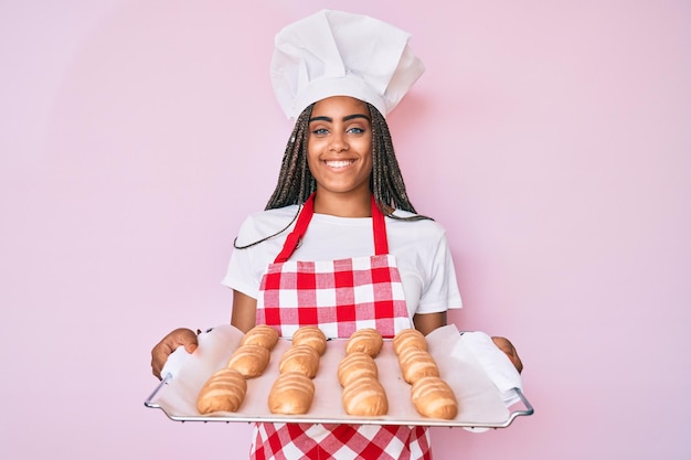 Foto grátis jovem mulher afro-americana com tranças vestindo uniforme de padeiro segurando pão caseiro sorrindo com um sorriso feliz e legal no rosto mostrando os dentes