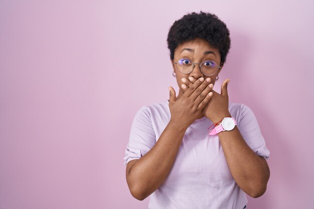 Jovem mulher afro-americana em pé sobre fundo rosa chocado cobrindo a boca com as mãos por engano. conceito secreto.