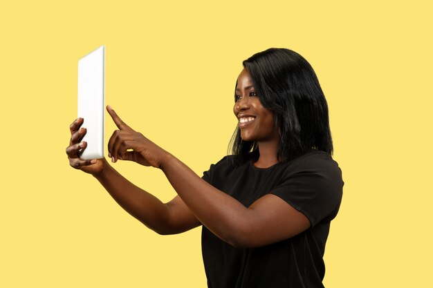 Foto grátis jovem mulher afro-americana isolada no fundo amarelo do estúdio, expressão facial. belo retrato feminino. conceito de emoções humanas, expressão facial. usando tablet para selfie ou vlog.