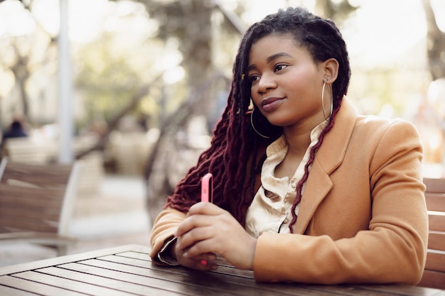 Jovem mulher afro-americana sentada em um café de rua segurando um smartphone nas mãos.