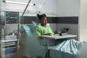 Foto grátis jovem mulher afro-americana usando tablet na cama da enfermaria de hospital. paciente doente com tecnologia online se recuperando de uma doença na clínica com equipamento médico. pessoa a navegar na web