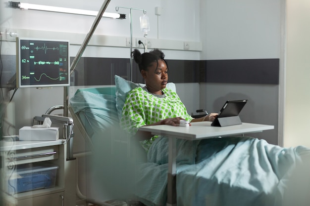 Jovem mulher afro-americana usando tablet na cama da enfermaria de hospital. Paciente doente com tecnologia online se recuperando de uma doença na clínica com equipamento médico. Pessoa a navegar na web