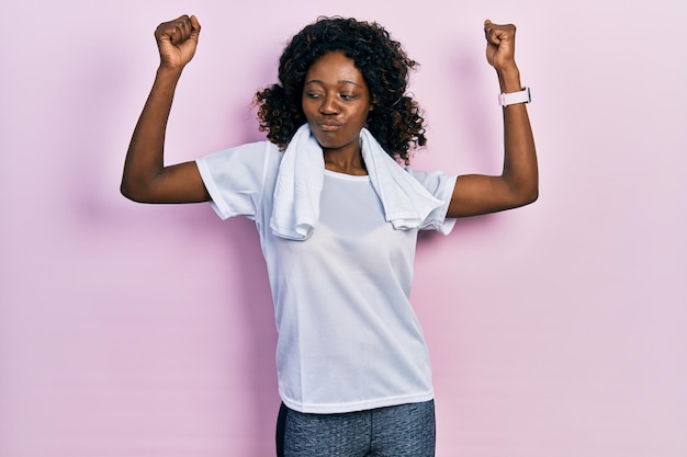 Jovem mulher afro-americana vestindo roupas esportivas e toalha mostrando os músculos dos braços sorrindo orgulhosos. conceito de aptidão.