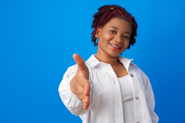 Jovem mulher afro esticando a mão para um aperto de mão contra um fundo azul