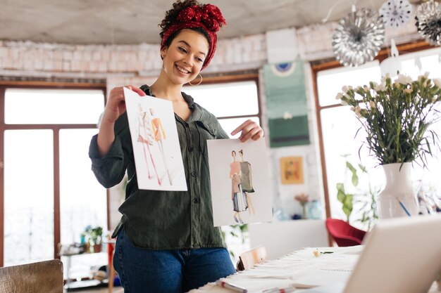 Jovem mulher alegre com cabelos cacheados escuros em pé perto da mesa mostrando alegremente esboços de moda no laptop enquanto passa o tempo na oficina moderna e aconchegante com grandes janelas