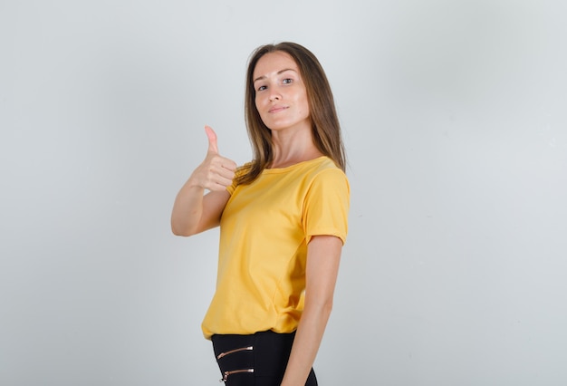 Jovem mulher aparecendo o polegar em uma camiseta amarela, calça preta e parecendo satisfeito.